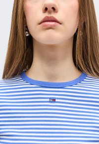 Νεαρό άτομο με μακριά ανοιχτοκάστανα μαλλιά, που φοράει μπλε και άσπρη ριγέ μπλούζα με μικρό λογότυπο στο κέντρο του στήθους και ασημένια σκουλαρίκια κρίκους.