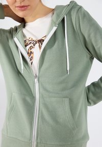 Sudadera de cremallera verde claro con una textura suave, cordones blancos, bolsillos laterales y cremallera completa. Lleva encima una camiseta de patrón.