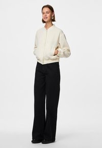 Žena nosi kremu bomber jaknu s elastičnim manšetama i strukom, uparenu s crnim širokim hlačama visokog struka i crnim cipelama.