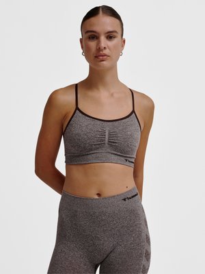 Vrouw staat met armen achter de rug, draagt een grijze naadloze sportbeha en bijpassende legging met zwarte bies en logo's op een effen achtergrond.