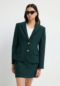 Mujer lleva un blazer verde oscuro con botones dorados, falda a juego, blusa blanca y gafas de sol negras tipo gato frente a un fondo liso.