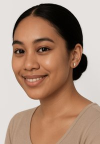 Silverfärgade studs i rund form med subtil textur, bärs av en person med mörkt hår och en ljusbrun topp, leende mot en neutral bakgrund.