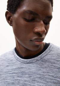 Grau gestrickter Pullover mit einem strukturierten Muster, runder Ausschnitt und schwarzem Saum. Nahaufnahme auf die Schulter und das Gesicht eines Modells.