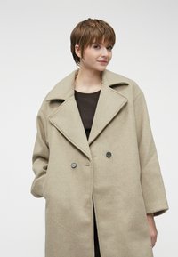 Μπεζ παλτό από μείγμα μαλλιού με διπλό κούμπωμα, φαρδύ κοψίμα για το κολάρο και κουμπιά κλεισίματος; διαθέτει χαλαρούς ώμους και άνετη εφαρμογή.