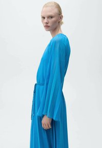 Rochie cu mânecă lungă, până la coapse, dintr-un material turcoaz transparent, cu pliseuri verticale, având o talie legată și o siluetă netedă, fluidă.