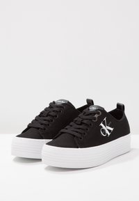 Baskets en toile noire avec une semelle plateforme blanche, des lacets et un logo sur le côté. Texture lisse et design à bout arrondi.