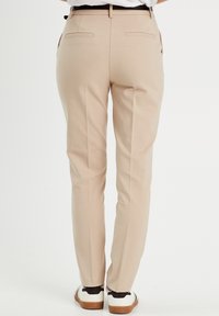 Pantalon beige sur mesure, coupe slim, avec deux poches arrière et une texture lisse, assorti à des baskets blanches avec des accents noirs.