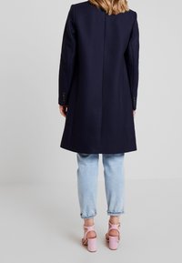 Abrigo de lana azul marino con corte recto, mangas largas y botones sutiles, combinado con jeans azul claro y sandalias de tacón rosas.