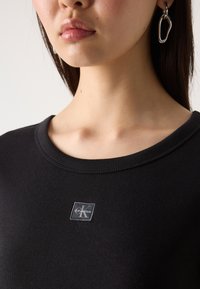 Black ribbed T-shirt with a crew neckline, featuring a small gray Calvin Klein logo patch at the chest. Smooth fabric texture.