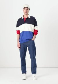 Farveblokeret pullover i marineblå, hvid, rød og blå, parret med blå jeans og hvide sneakers, suppleret med en beige kasket.