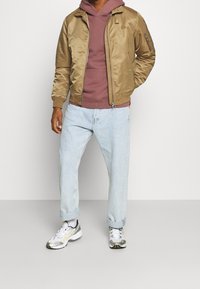 Brązowa satynowa kurtka bomber na ciemnoczerwonej bluzie z kapturem, w połączeniu z jasnoniebieskimi dżinsami i białymi trampkami z szarymi i żółtymi akcentami.