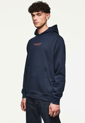 Młody mężczyzna z kręconymi włosami, ubrany w granatowy hoodie z napisem "WEEKEND OFFENDER ADMIT NOTHING", stojący na jednolitym jasnym tle.