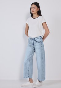 T-shirt blanche à manches courtes avec encolure noire, associée à un jean large bleu clair. Le mannequin porte des baskets blanches et se tient devant un fond uni.