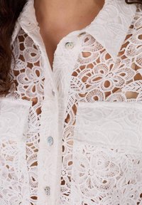 Witte kanten blouse met knoopsluiting, voorzien van bloemmotieven, twee voorzakken en lichtblauwe knopen op een semi-transparante stof.