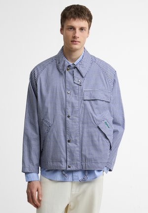 Noor mees, kes kannab sinise ja valge ruudulise jaki all helerohelist särki ja beežid pükse, seisab ühevärvilise valge tausta ees.