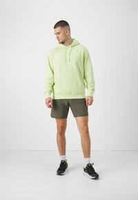 Felpa verde neon con tasca frontale, cordini e logo ricamato. Abbinata a pantaloni corti verdi oliva e scarpe da ginnastica nere con dettagli neon.