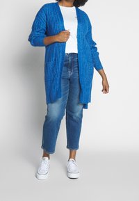 Cardigan bleu côtelé avec poches latérales, porté sur un t-shirt blanc, associé à un jean bleu et des baskets blanches. Texture douce, coupe décontractée.