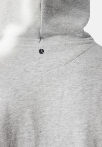 Gaiši pelēka sporta jaka, kas fokusējas uz augšējo muguras daļu un kapuci ar mazu melnu izšūtu logotipu zem kapuces šuves.