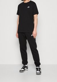 Person som bär svart Nike T-shirt och joggingbyxor med vita Nike-logotyper, kombinerat med svartvita sneakers, stående mot en enfärgad bakgrund.