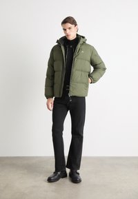 Olivgrüne Pufferjacke mit Kapuze, ausgestattet mit einem Reißverschluss, elastischen Bündchen und einem Logo-Patch auf der Brust, getragen über einem schwarzen Pullover.