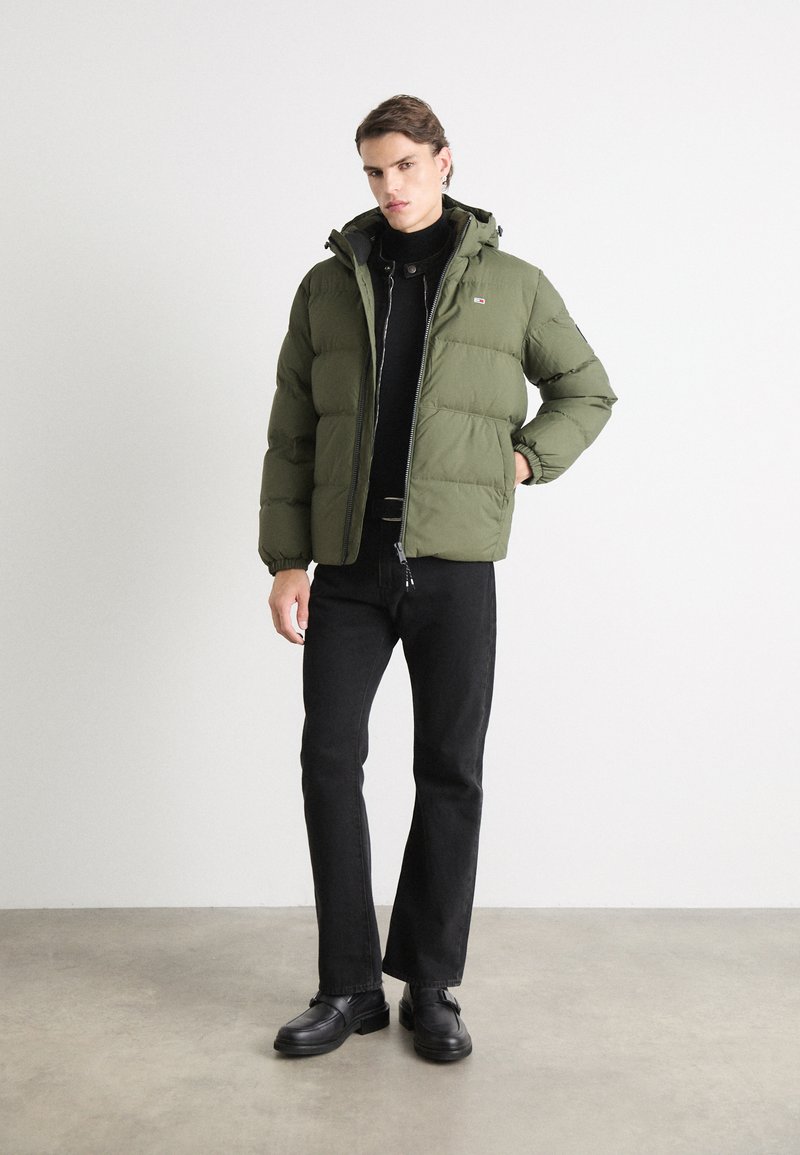 Olivgrüne Pufferjacke mit Kapuze, ausgestattet mit einem Reißverschluss, elastischen Bündchen und einem Logo-Patch auf der Brust, getragen über einem schwarzen Pullover.
