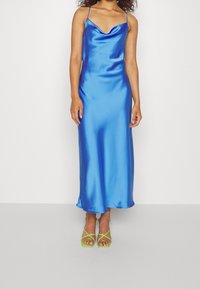 Blue satin slip dress with a cowl neckline, thin straps, and a flowing silhouette, paired with green strappy heels.