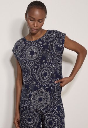 Kobieta z zaplecionymi włosami, ubrana w granatowy top i spodnie z białymi, misternymi, okrągłymi wzorami, pozująca z jedną ręką na biodrze.