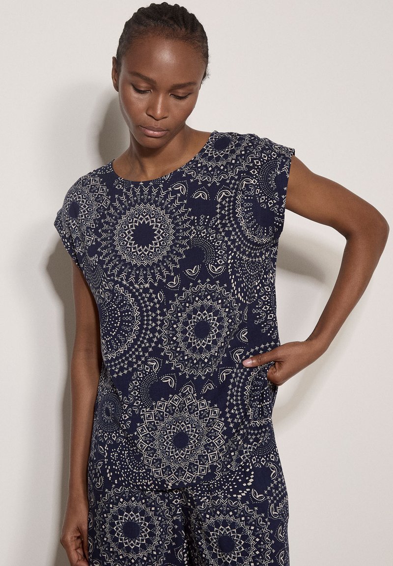 Femme aux cheveux tressés portant un haut et un pantalon bleu marine avec des motifs circulaires blancs détaillés, posant avec une main sur la hanche.