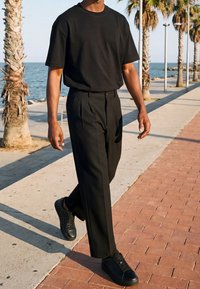 Homme vêtu d'un t-shirt noir, pantalon et chaussures, marchant sur un chemin pavé près de palmiers au bord de la mer par une journée ensoleillée.