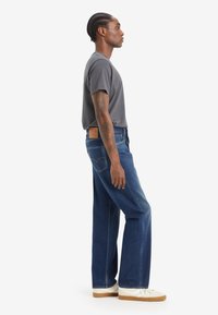 Maglietta grigia a maniche corte abbinata a jeans azzurri a vita alta con una toppa in pelle marrone. Sneakers bianche con suole beige completano il look.