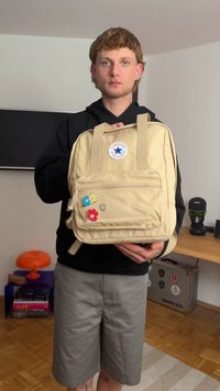 Világosbarna vászontáska, tetején fogantyúval, állítható pántokkal, elülső zsebbel, csillag logóval ellátott folttal és színes virágformájú díszítésekkel.