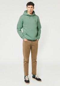 Green hooded sweatshirt with a front pocket, paired with tan corduroy pants and black sneakers featuring white laces and green accents.