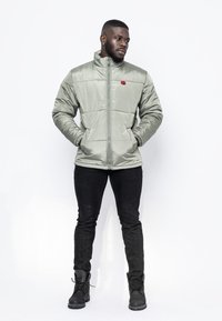 Vadderad ljusgrön jacka med hög krage, framdragkedja och röd logotyp; bärs över svarta slim-fit byxor och svarta snörboots.