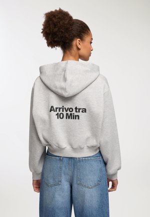 Donna con capelli ricci che indossa una felpa grigia con il testo "Arrivo tra 10 Min" sulla schiena e jeans di denim blu, di spalle contro uno sfondo bianco.