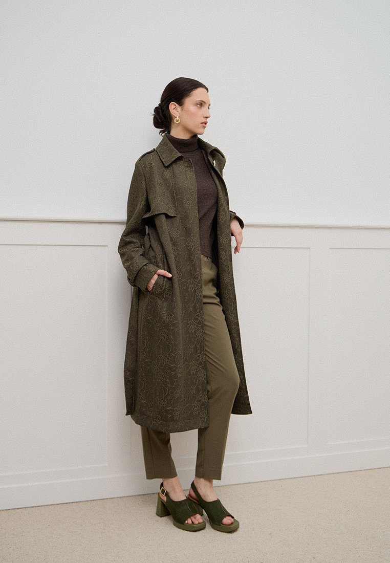 Lungo trench verde oliva con un motivo texturizzato, abbinato a un dolcevita marrone scuro e pantaloni oliva, e sandali con tacco verdi.