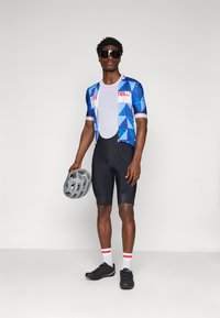 Tenue de cyclisme comprenant un maillot bleu à motifs géométriques, un short noir, des chaussures de cyclisme noires, des chaussettes blanches avec des rayures rouges, et un casque gris.