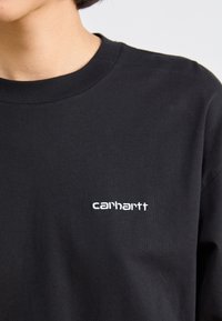 T-shirt en coton noir avec un col rond, présentant un petit logo "carhartt" brodé en blanc sur la poitrine gauche. Texture lisse.