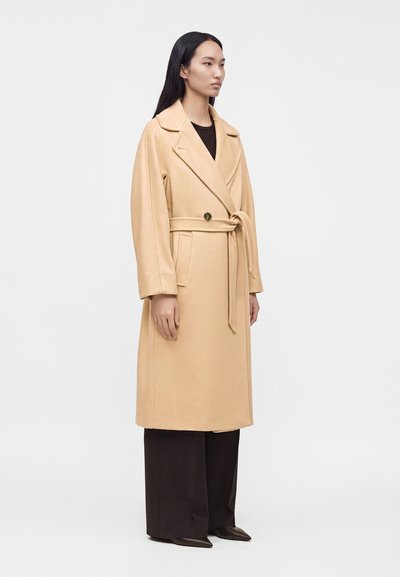 Femme debout de profil, vêtue d’un long manteau beige ceinturé par-dessus un pantalon noir à jambes larges et des chaussures noires pointues sur un fond uni.