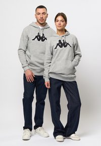 Szürke kapucnis pulcsik fekete grafikus logóval, kenguruzsebekkel és húzózsinórokkal. Sötét farmernadrággal és fehér sneaker cipővel párosítva.