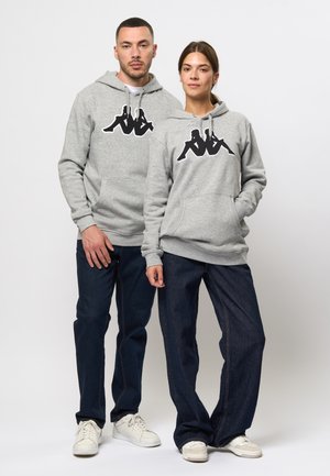 Grijze hoodie met een zwart grafisch logo, met kangoeroezakken en koordjes. Gepaard met donkerdenim jeans en witte sneakers.