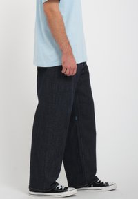 Pantalones de mezclilla oscuros de pierna ancha con una textura suave, que presentan un detalle del logo en azul, llevados con zapatillas negras de corte bajo y una camisa de color azul claro.