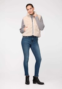 Beige mākslīgās kažokādas veste ar augstu apkakli, rāvējslēdzēja aizdari un pelēkiem adījuma piedurknēm, apvienota ar ziliem pieguļošiem džinsiem un melnām potīšu zābakiem.