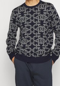 Maglione blu navy con motivo geometrico a catena grigia, collo rotondo, polsini e fondo a coste, texture lavorata a maglia e design attillato.