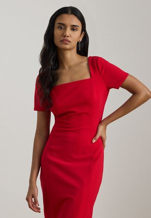 Frau mit langen dunklen Haaren, die ein eng anliegendes rotes Kleid mit kurzen Ärmeln und quadratischem Ausschnitt trägt, steht mit einer Hand in der Hüfte.