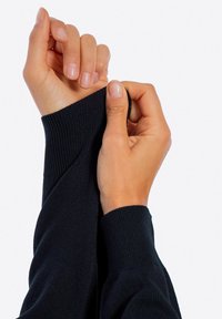 Czarny dzianinowy sweter z prążkowanymi mankietami; ręce ciągnące za rękaw. Gładka faktura, dopasowany krój. Skupienie na detalu mankietu.