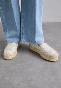 Pārklāta bežā espadrilles ar austu jutu zoli, ar gaiši brūnu akcentu purngala daļā un smalku ģeometrisku rakstu augšējā daļā.