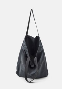 Bolsa de tela de color gris oscuro con una larga correa para el hombro y asas más cortas, mostrada abierta contra un fondo claro y liso.