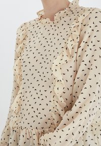 Blusa crema con motivi a forma di cuore neri, colletto e maniche volanti, tessuto testurizzato e vestibilità ampia con dettagli arricciati.