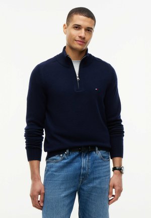 Homme debout portant un pull bleu marine à fermeture éclair semi-ouverte, un sous-vêtement gris clair, un jean bleu, une ceinture noire et une montre noire sur fond blanc.