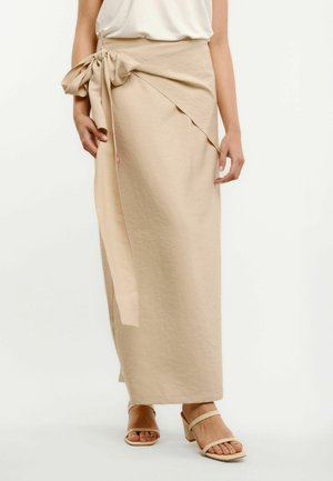 Bézs, átkötős midi szoknya nagy, bal csípőn megkötött masnival, fehér felsővel és bézs, nyitott orrú, vastag sarkú szandállal viselve.